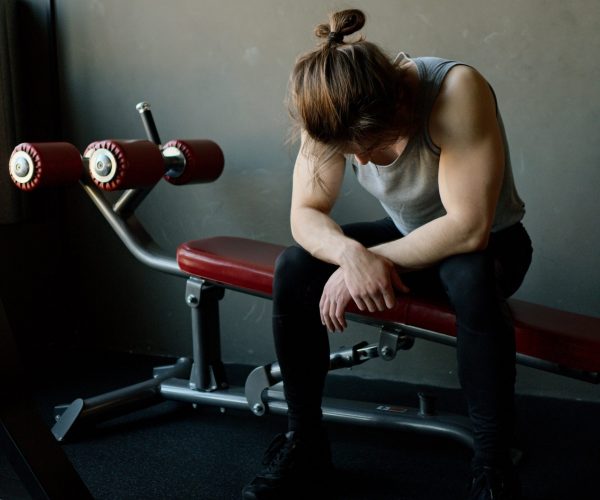 January-Fitness-Business-Slump A man sits on a bench in a gym. He's slumped over and holds a sweaty towel in his hands.